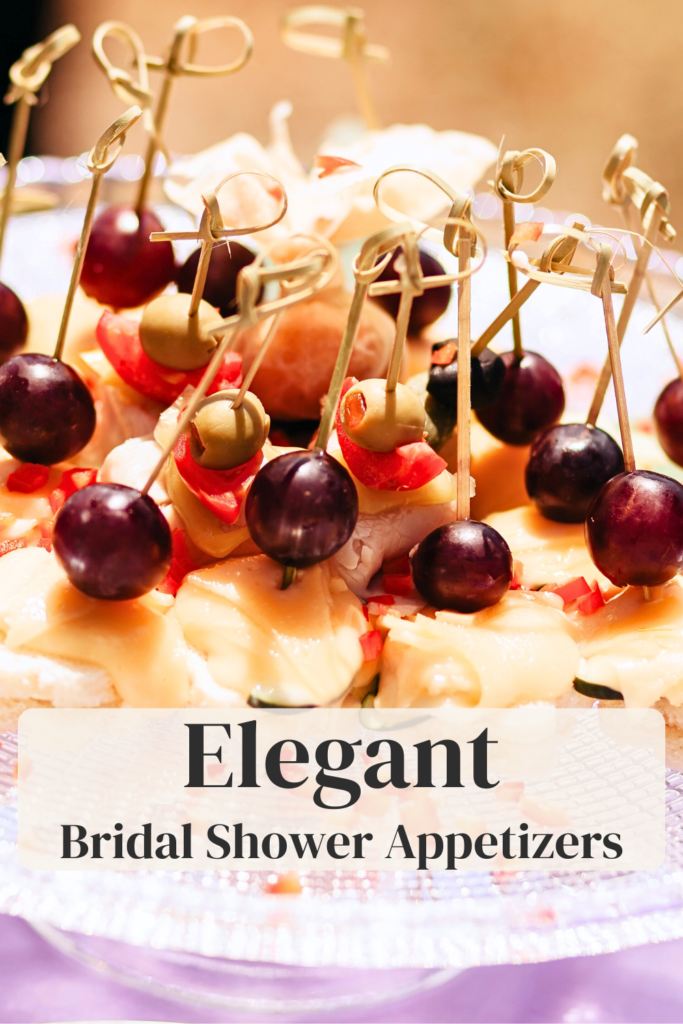 Elegant bridal shower appetizers arranged on a glass stand with brie, grapes, olives, and red bell pepper on mini bread slices.